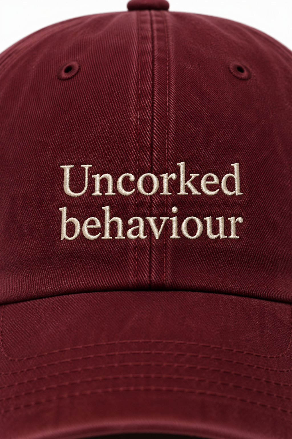 Close up maroon burgandy cap with 'Uncorked behaviour' embroidered text against a white background