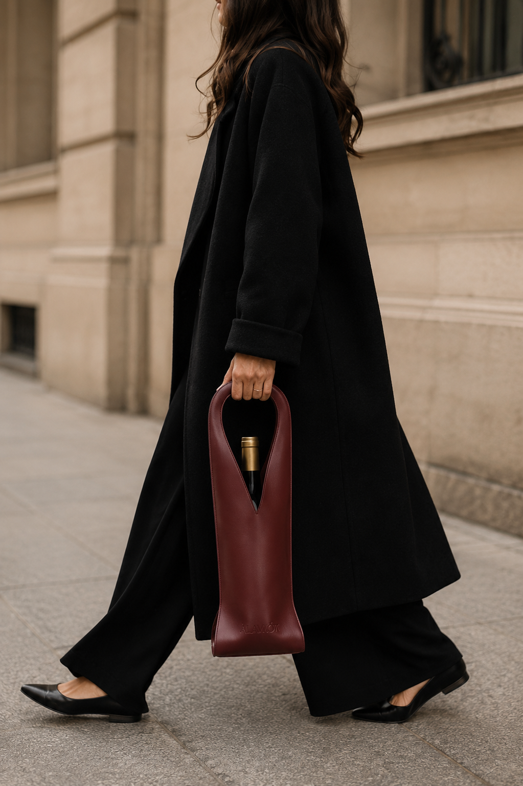 Person in a long black coat holding a burgundy tote bag with a bottle inside, walking on a sidewalk.