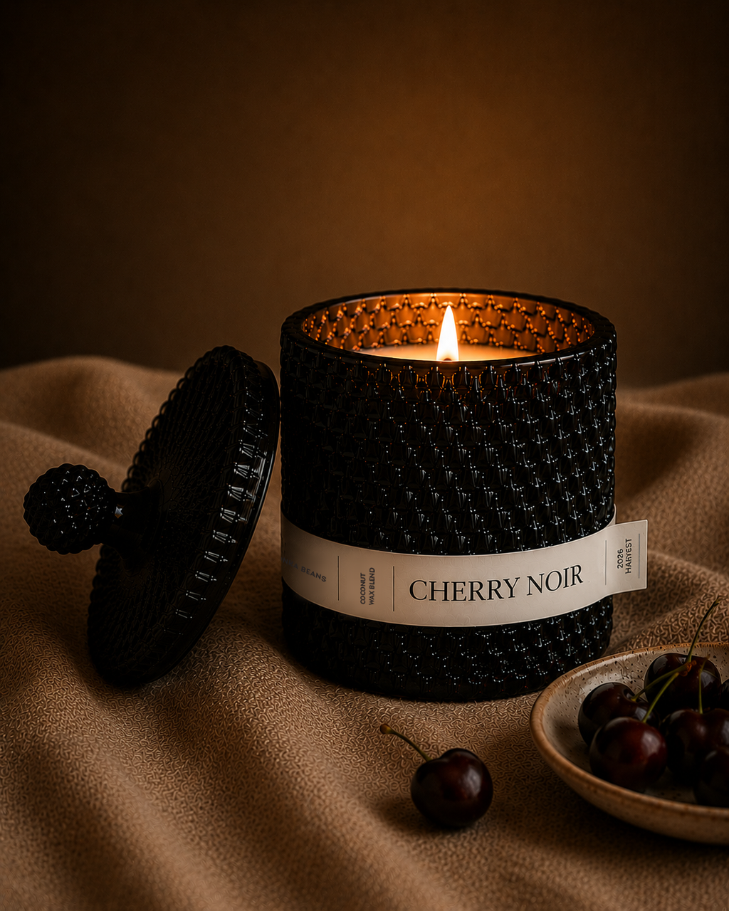 Black candle labeled 'Cherry Noir' with a lit wick on a brown fabric background.