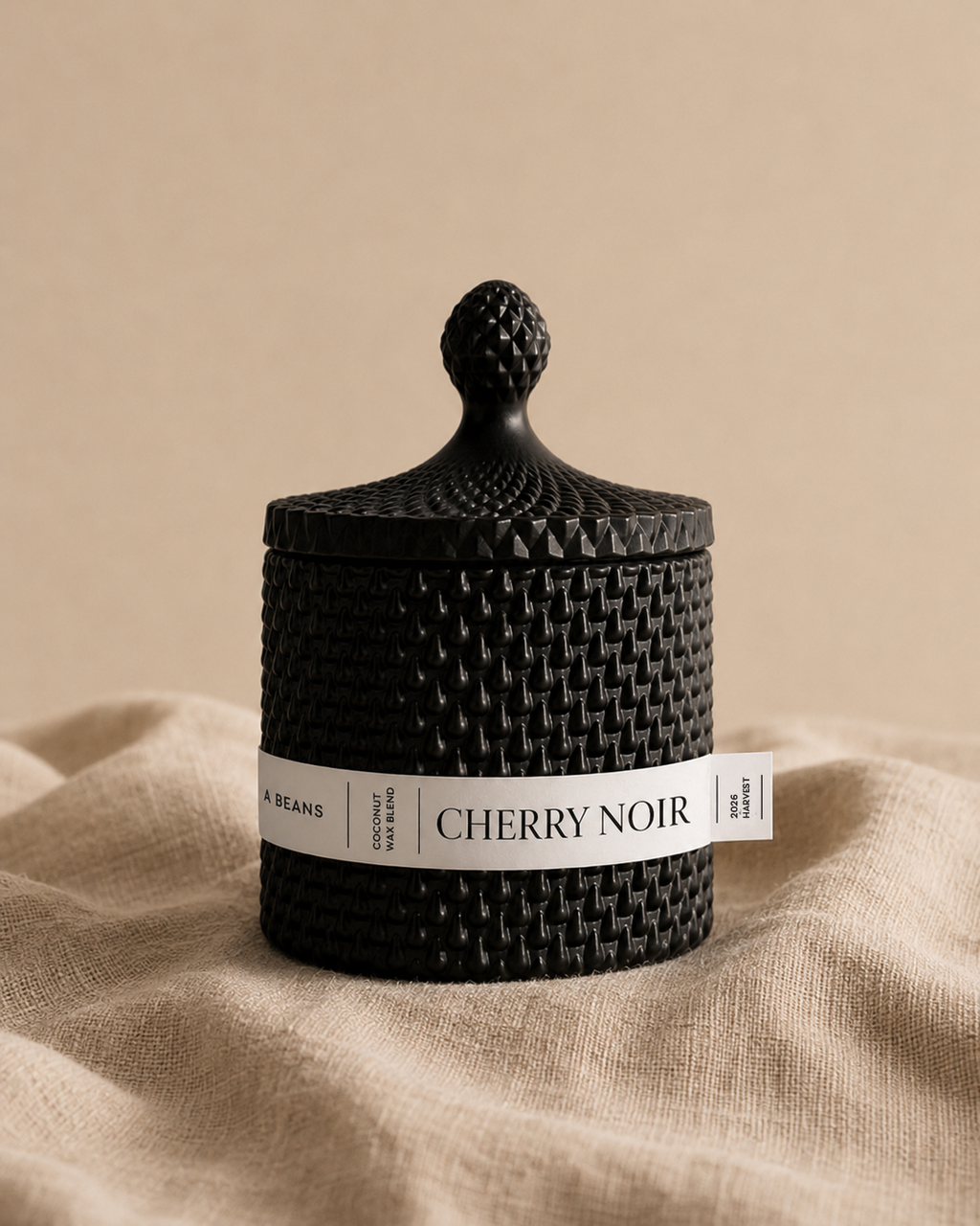 Black scented candle labeled 'Cherry Noir' on a beige fabric background