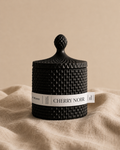 Black scented candle labeled 'Cherry Noir' on a beige fabric background