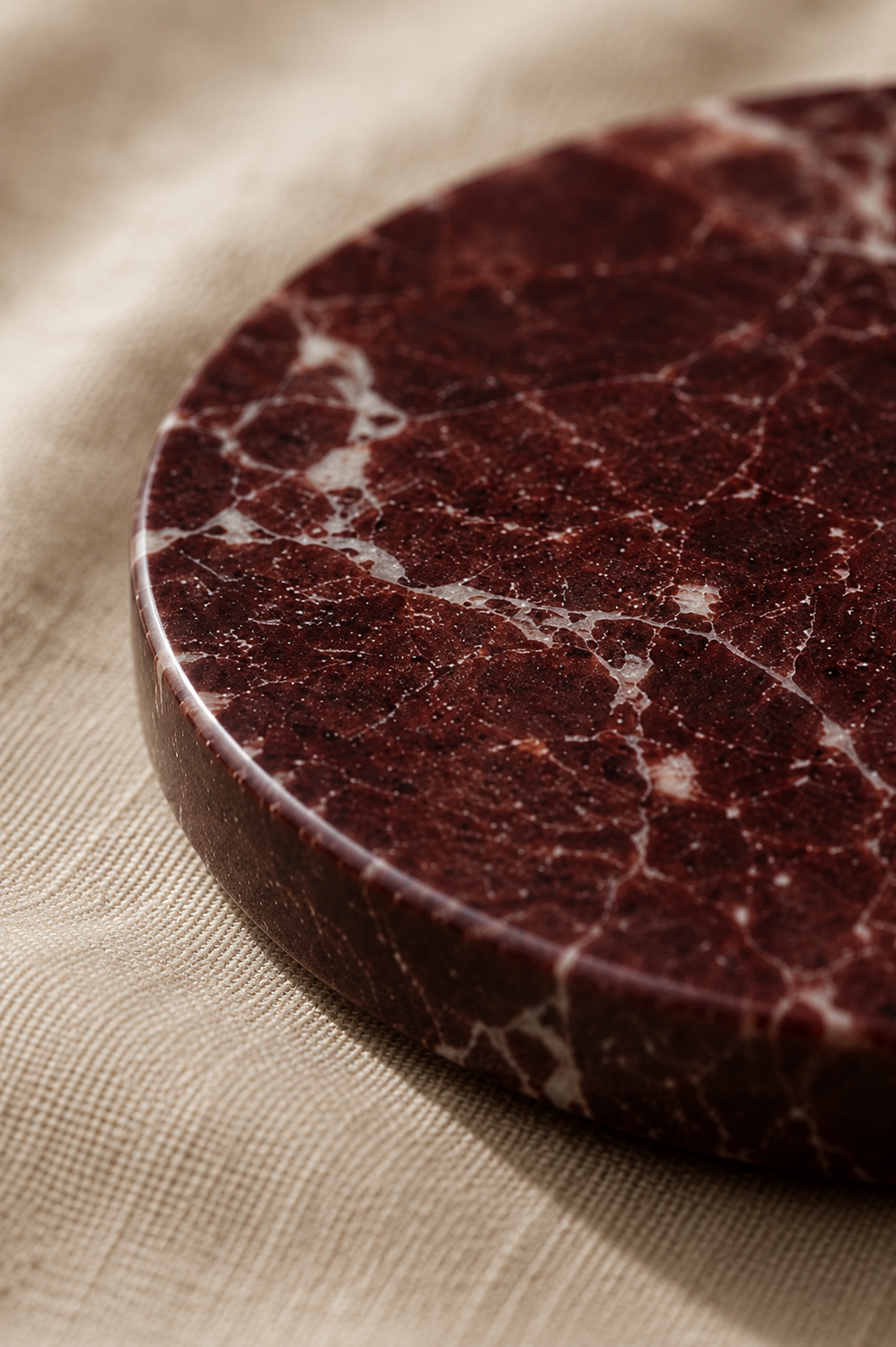 Close-up of a round, dark red stone marble drink wine coaster on a beige fabric background