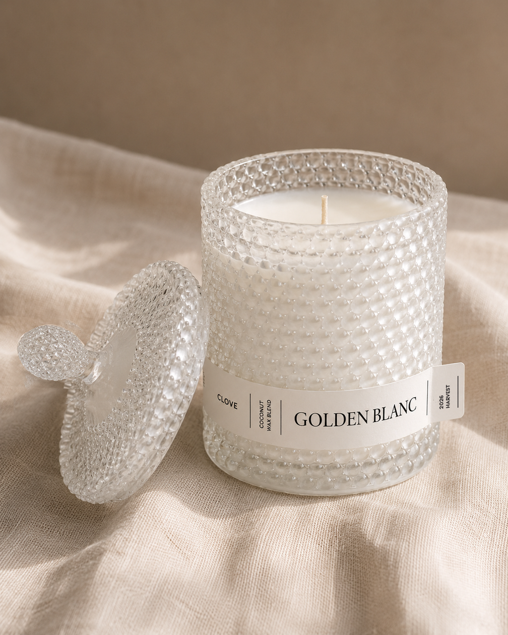 Decorative scented candle with a textured white lid and label on a linen table cloth. Alavot